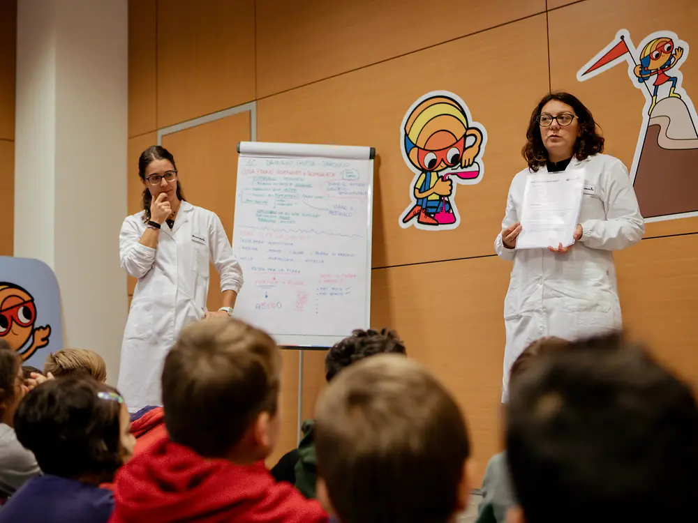 Two adults in lab coats stand in front of a group of children. One person is presenting a sheet of paper, while the other stands next to a flipchart with notes. The room is decorated with Researchers’ World illustrations.