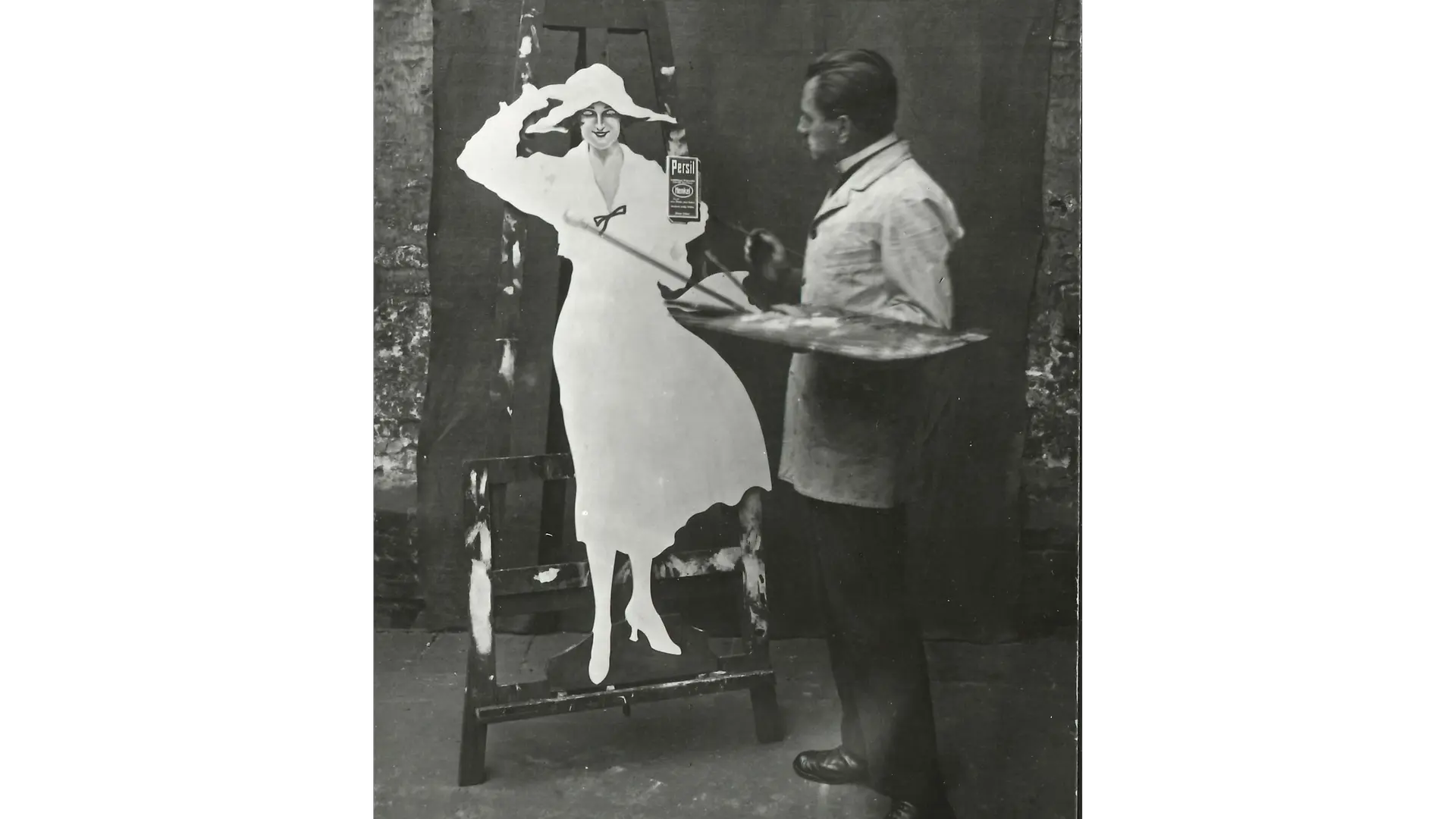 A black‑and‑white historical photograph, showing a person standing in front of a large board, painting or touching up the outline of Persil‘s Lady in White.