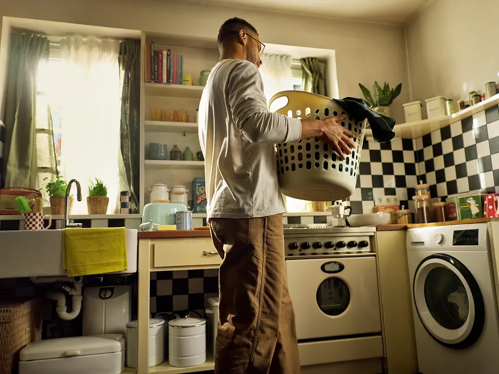 A man holding a large laundry basket filled with clothes is walking towards his washing maching in a cozy, retro‑style kitchen.