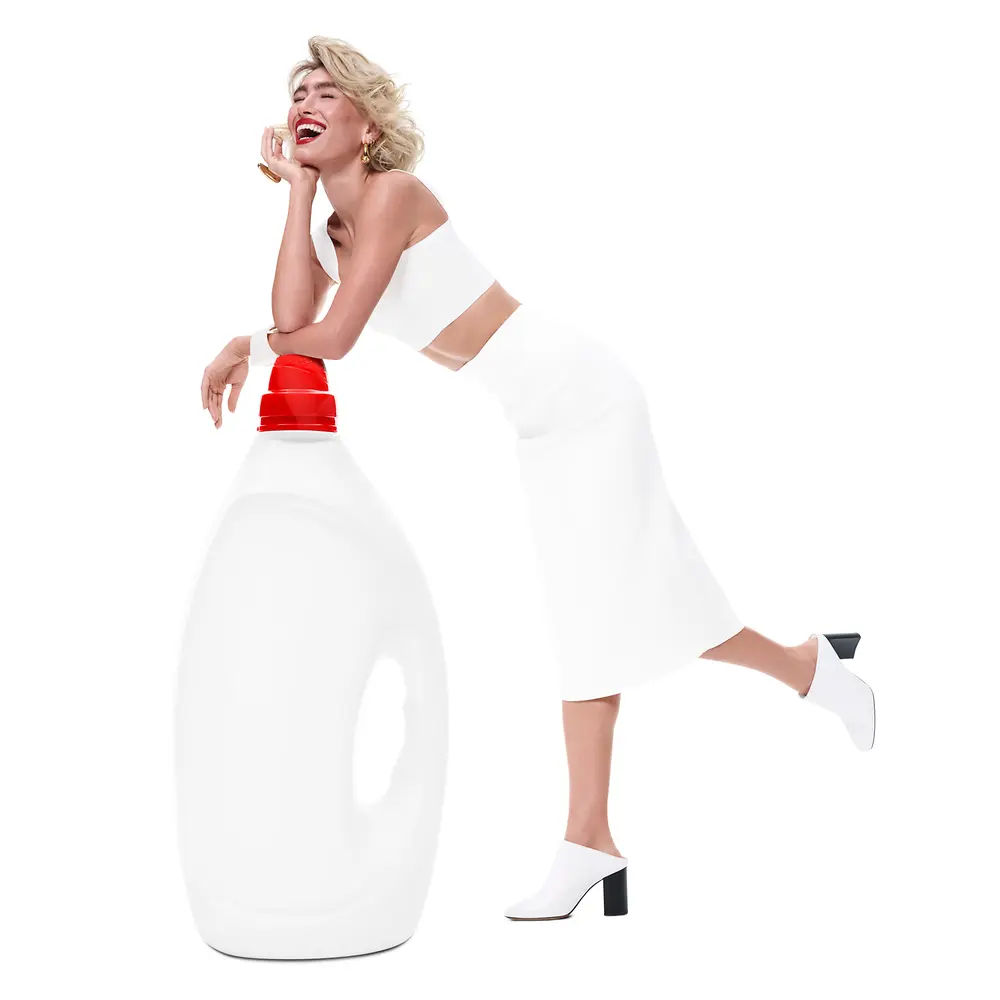 A woman dressed in white leans casually on an oversized white detergent bottle with a red cap, against a white background. The woman stands on one leg with the other bent back.