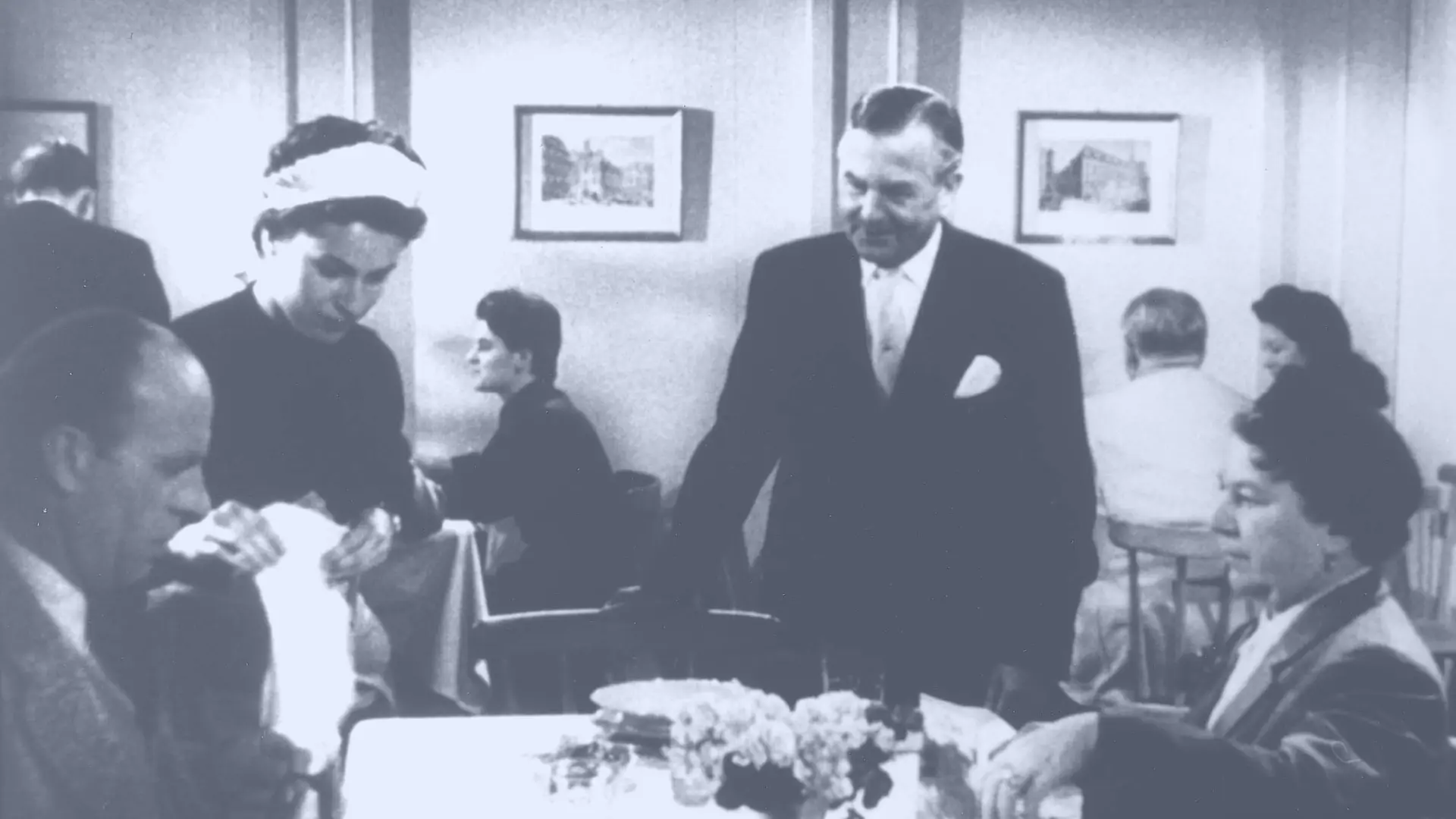 A black‑and‑white snapshot from the first Persil commercial showing several people seated or standing around a dining table in an indoor setting.