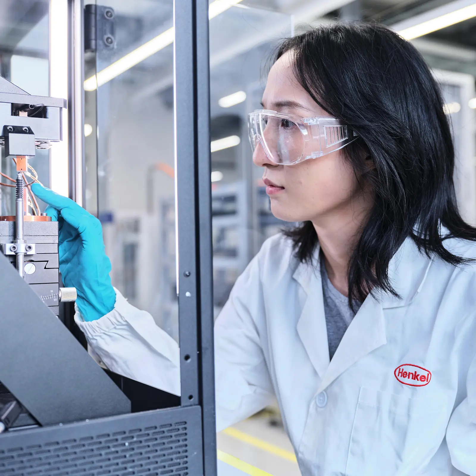 A young woman wearing a lab coat and safety glasses operates lab machinery.