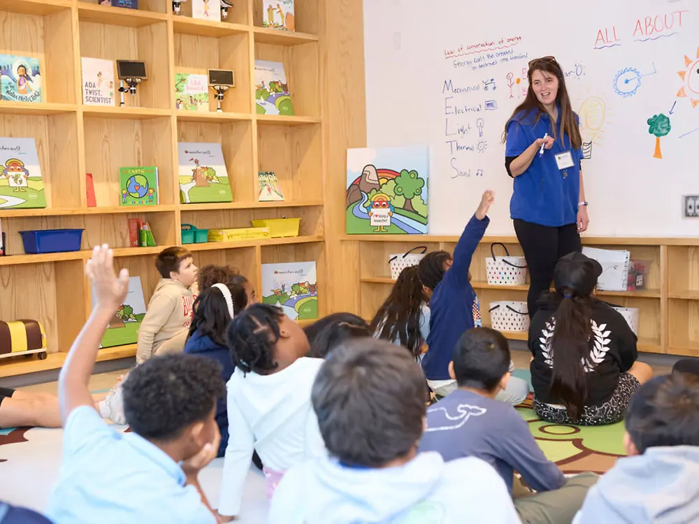 An educator is standing in front of a whiteboard, while several children are sitting on the floor behind her. Several of the children are raising their hand.