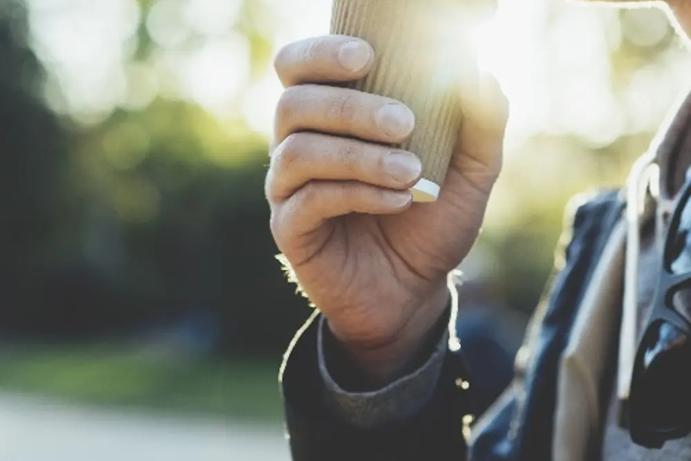 Close-up of a person holding a small takeaway coffee cup with a lid near the mouth, wearing a jacket. The background is softly blurred with trees and sunlight creating a warm, natural atmosphere.