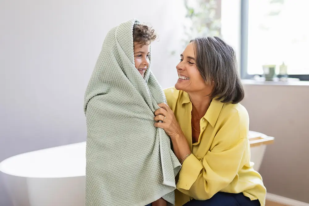 An adult gently wrapping a child in a textured towel in a bright bathroom setting. The child is standing near a bathtub while the adult, wearing a light-colored top, holds the towel around the child. Natural light enters through a window, illuminating a clean, minimal interior with soft, neutral tones.