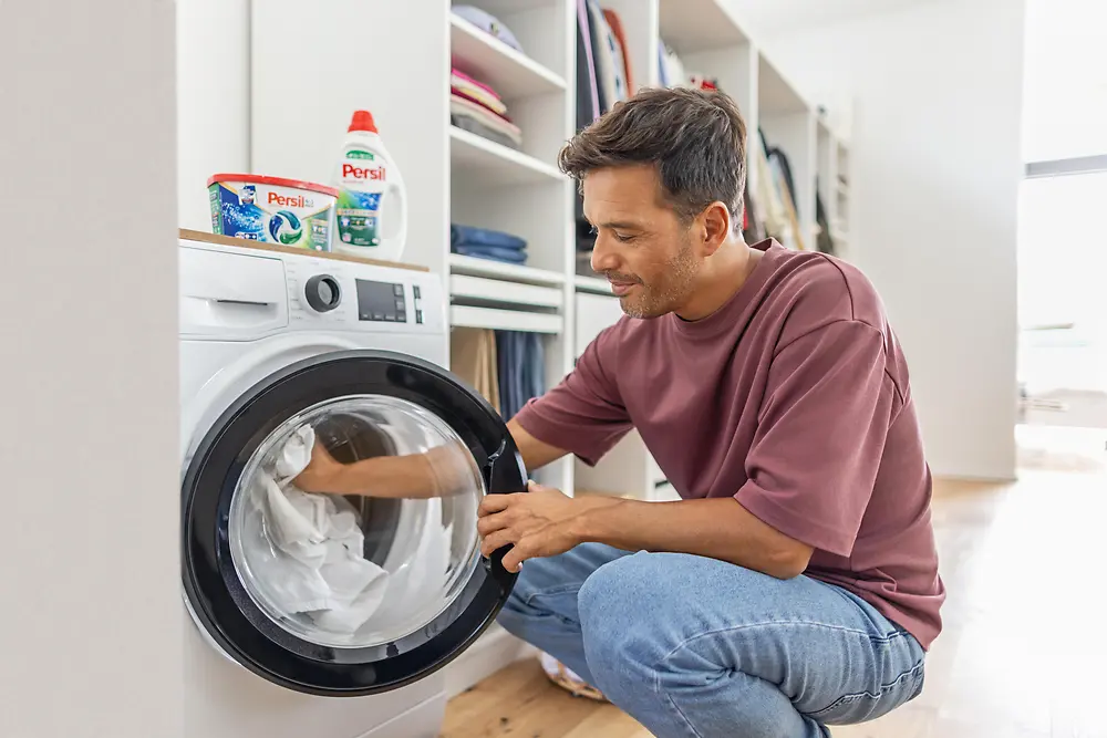A person crouching in front of a front-loading washing machine, placing clothes inside. Laundry detergent products are visible on top of the machine, and neatly arranged shelves with folded clothes are in the background of a bright, modern utility room.
