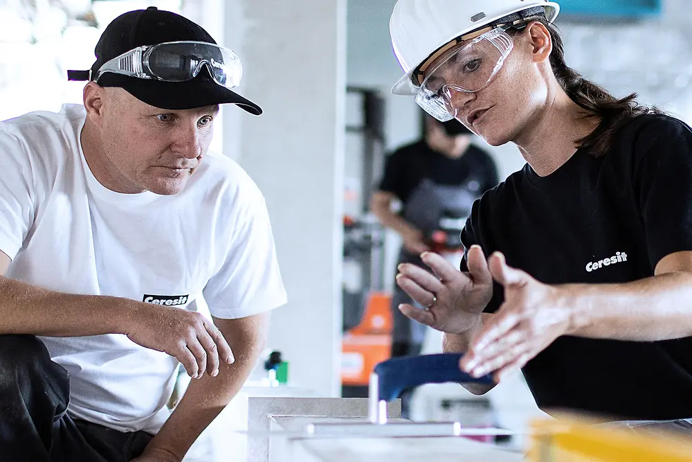 Two workers wearing protective gear, including safety glasses and a hard hat, kneel beside a partially finished surface indoors. They are discussing or inspecting a task, with tools and building materials nearby. In the background, another worker and a bright, industrial-style workspace are visible.