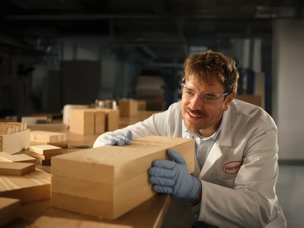 Person in a Henkel laboratory examining a rectangular wooden block on a workbench.