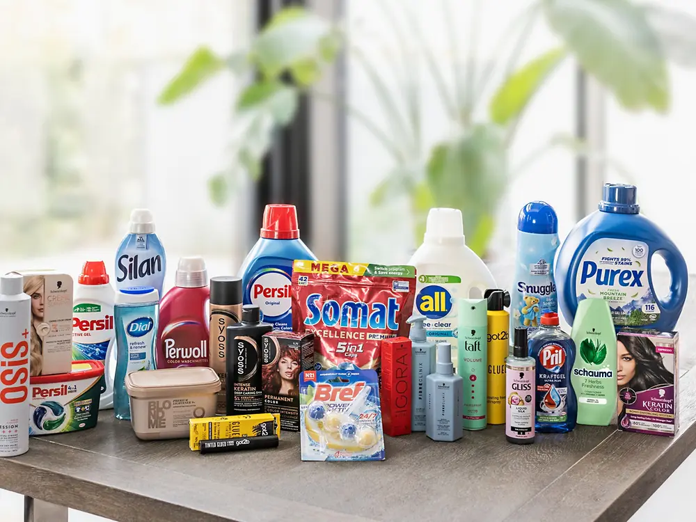 Ausgewählte Produkte aus dem Henkel-Portfolio stehen auf einer Tischplatte, darunter Marken aus den Bereichen Wasch- und Reinigungsmittel sowie Haarpflege und Styling. Die Anordnung verdeutlicht die Vielfalt der Consumer Brands von Henkel.