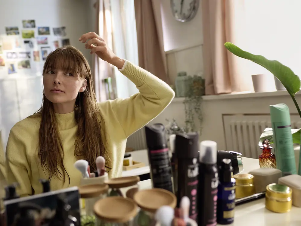 Young woman sitting in front of a mirror styling wet hair, with various hair care and styling products arranged on the countertop.