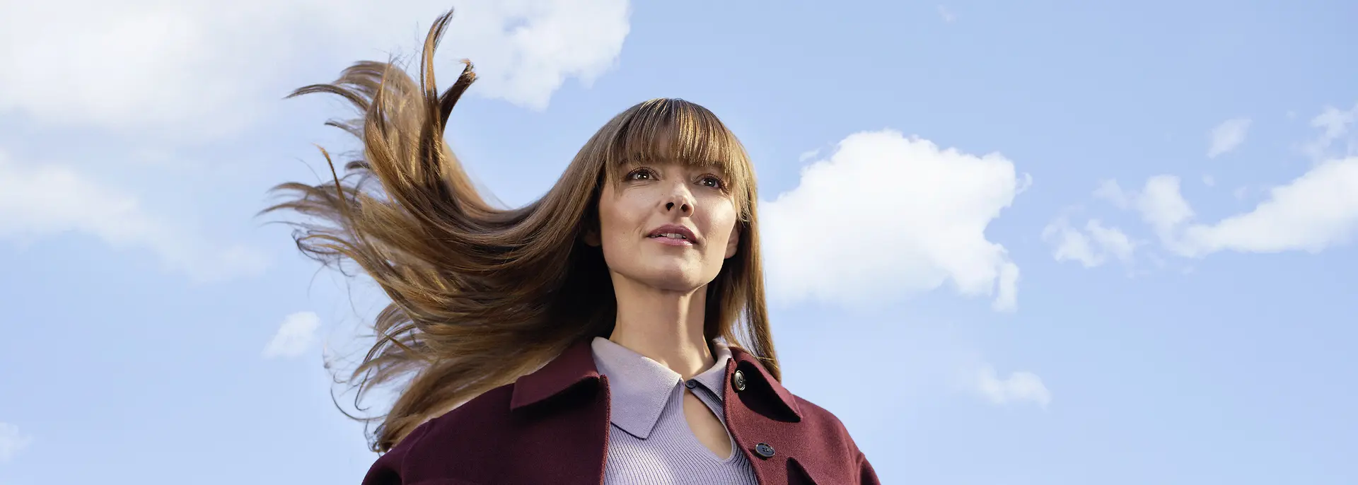 Young woman with long brown hair blowing in the wind against a blue sky with white clouds.
