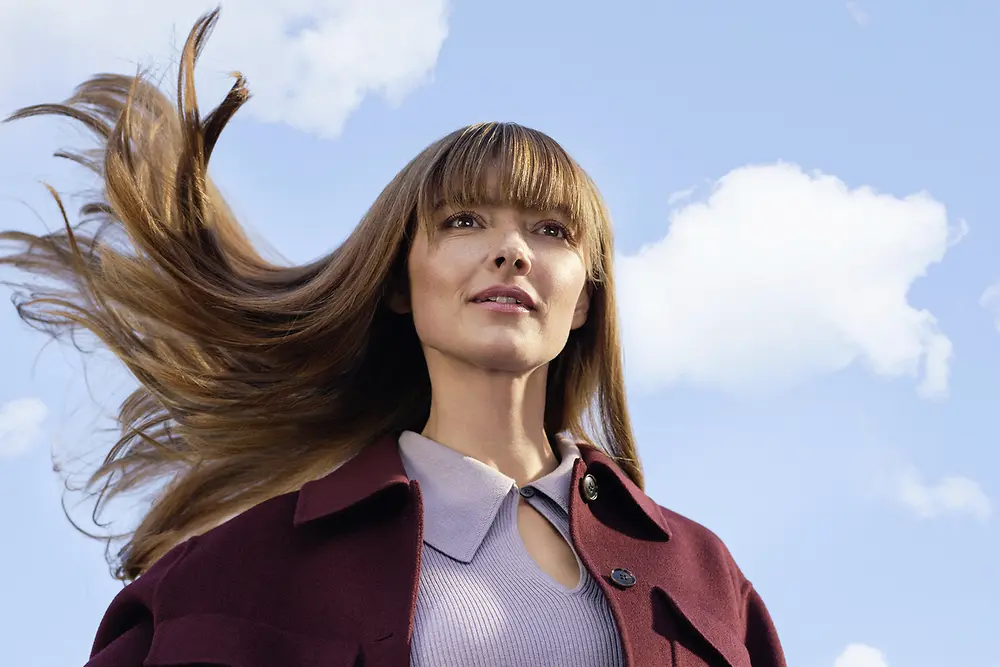 Young woman with long brown hair blowing in the wind against a blue sky with white clouds.