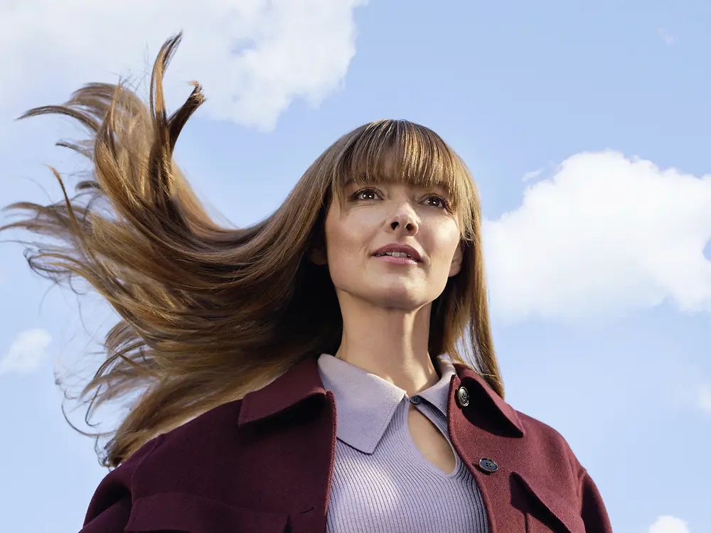 Young woman with long brown hair blowing in the wind against a blue sky with white clouds.
