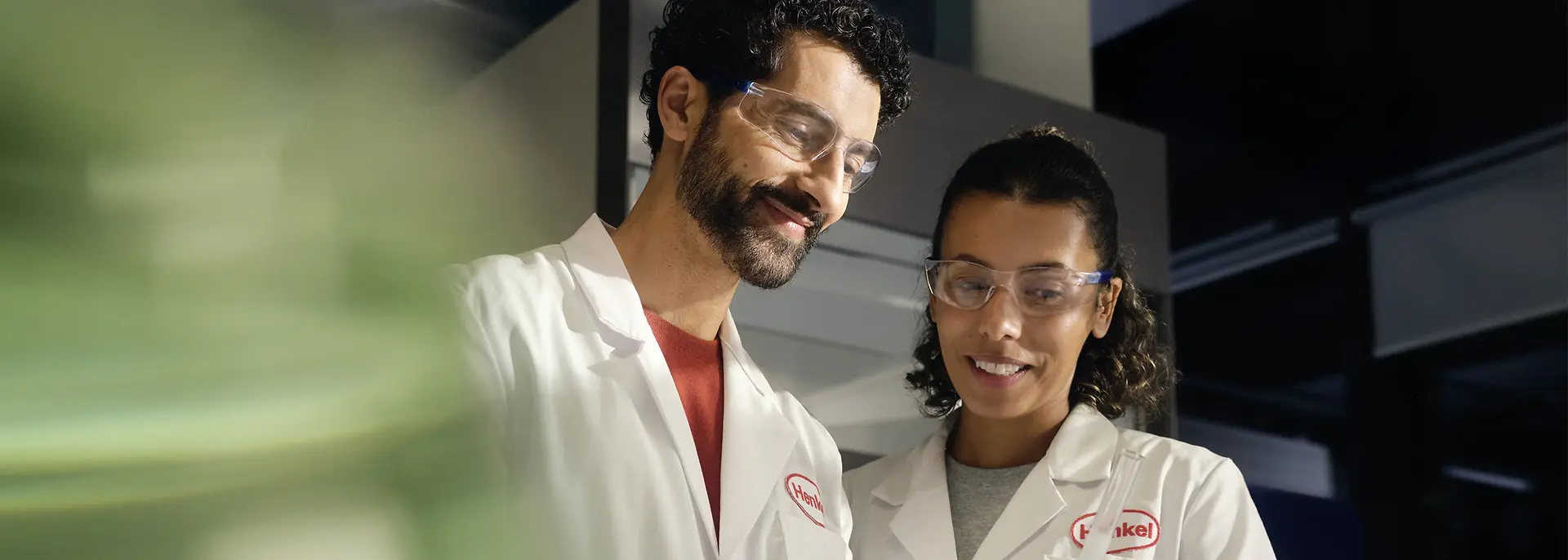 Two researchers in Henkel lab coats analyze a vial.