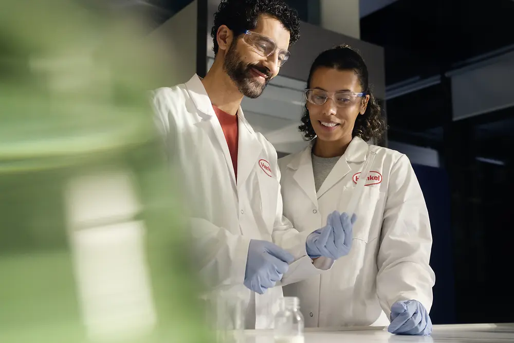 Two researchers in Henkel lab coats analyze a vial.