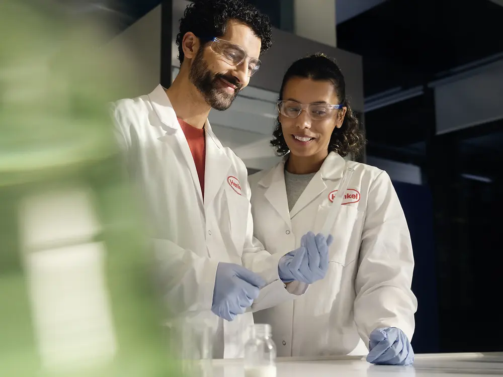 Two researchers in Henkel lab coats analyze a vial.