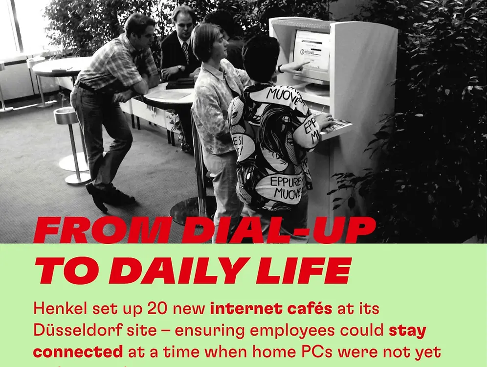 Black-and-white photo of several people gathered around a large early desktop computer in an indoor space. One person points at the monitor while others stand or lean nearby, looking at it. Tables, chairs, and plants appear in the background. Bold red text reads “FROM DIAL-UP TO DAILY LIFE.” Supporting text explains that Henkel set up 20 internet cafés at its Düsseldorf site so employees could stay connected when home PCs were not yet widespread. The year “2001” appears prominently at the bottom.