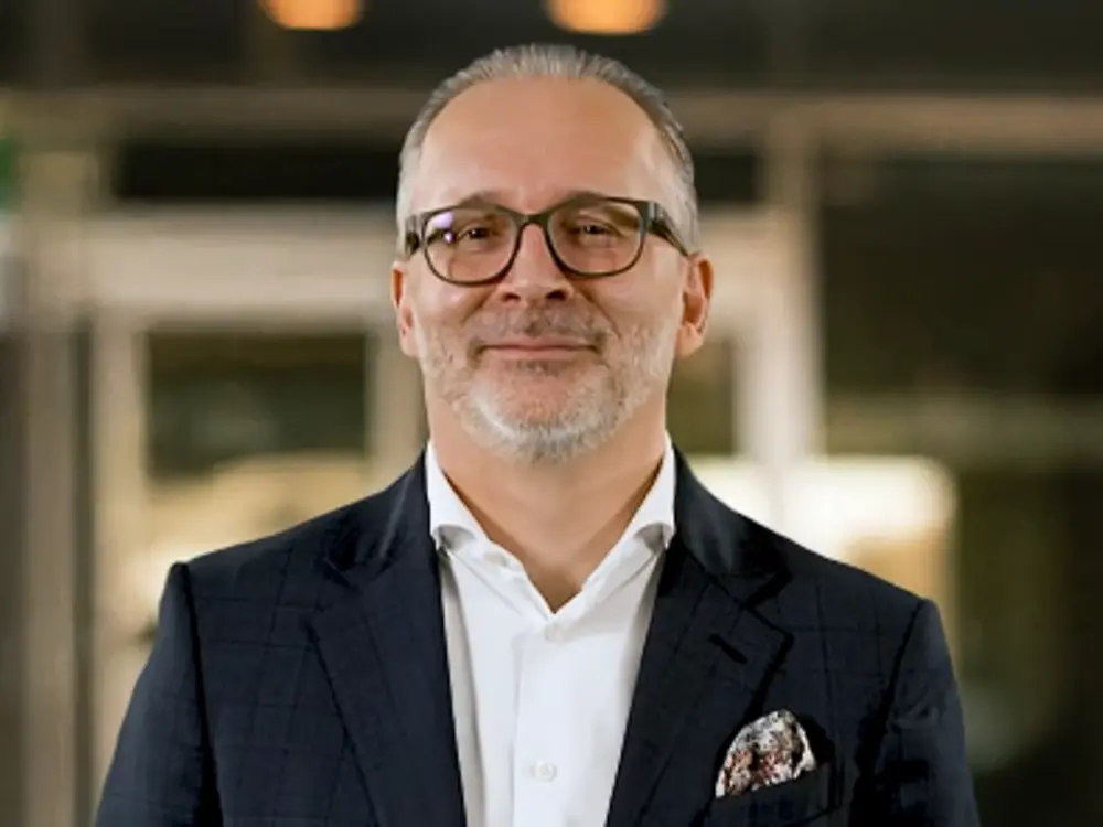 Portrait of a person wearing a dark suit and a white shirt. The person is standing with folded hands in front of a blurred indoor background and looking directly at the camera. 