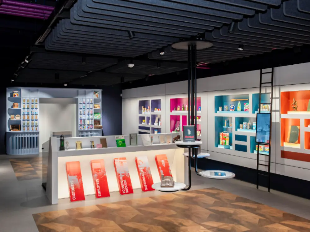 Interior view of a modern exhibition space featuring illuminated display walls with historical and current consumer products, colorful shelving units, interactive exhibits, and branded showcases. A central display island presents product-related artifacts and information panels, while contemporary lighting and architectural elements create a museum-like environment highlighting innovation and brand heritage.