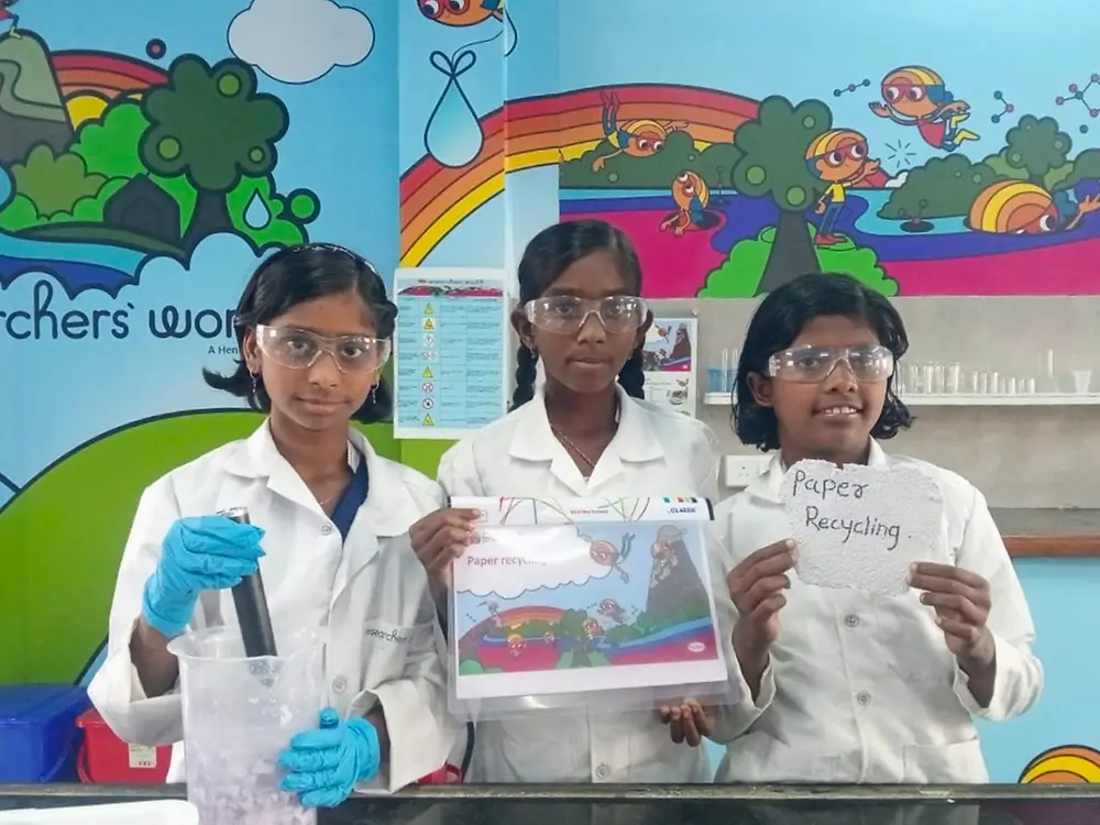 Three children wearing lab coats and safety glasses present the results of a paper-recycling experiment.