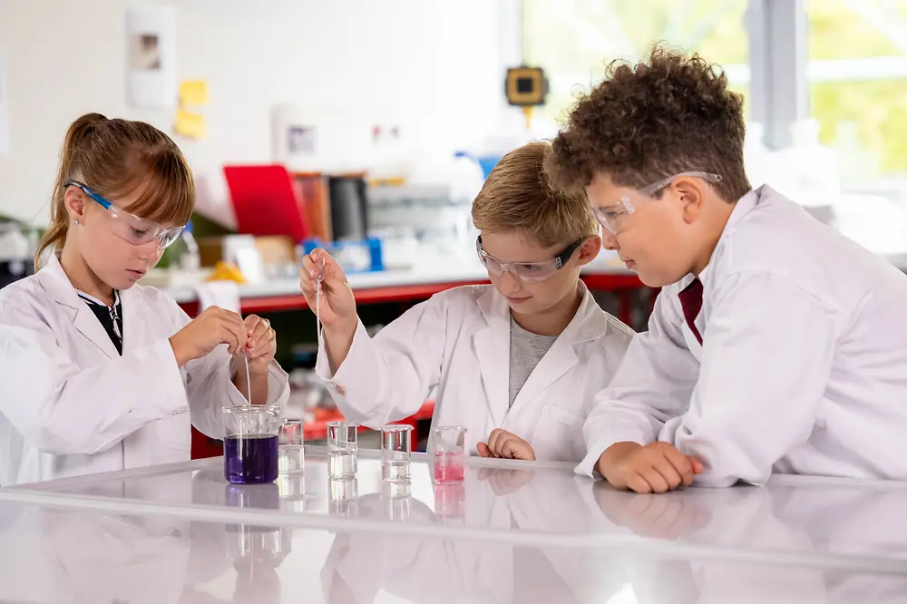 Three children wearing lab coats and safety glasses conduct an experiment with colored liquids.
