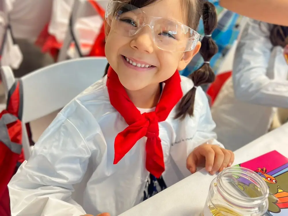 A smiling child wearing a lab coat and safety glasses sits at a table during a Researchers’ World activity, with an experiment setup in front of her.