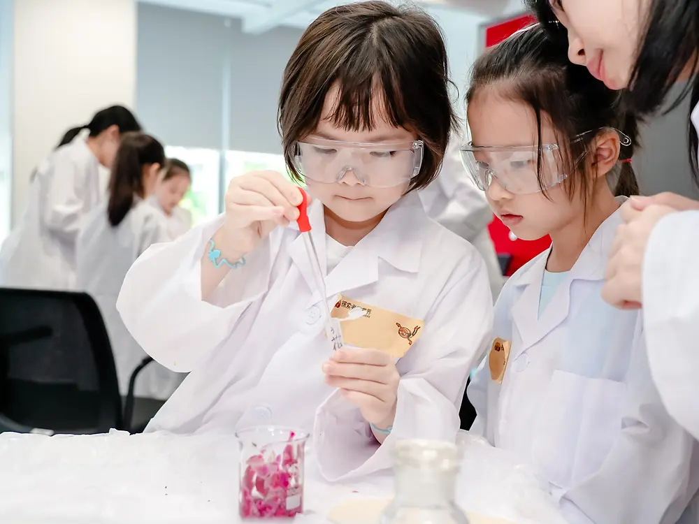 Two children wearing lab coats and safety glasses conduct an experiment with cups and pipettes under supervision.