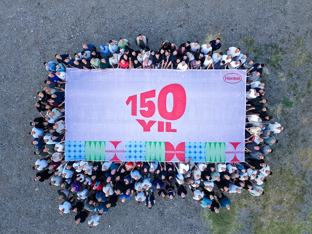 A large group of employees stands around a banner displaying the Henkel 150 Years anniversary logo, forming a group image from above.