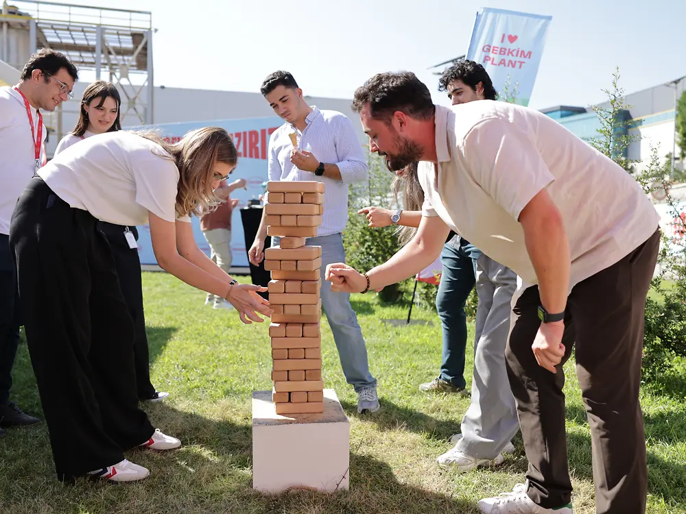 Several employees play a giant Jenga game outdoors, carefully removing wooden blocks from a tall tower.