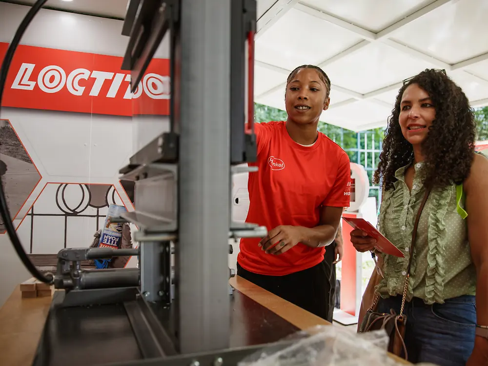 A Henkel employee demonstrates a Loctite technology exhibit while a visitor observes the application and equipment.