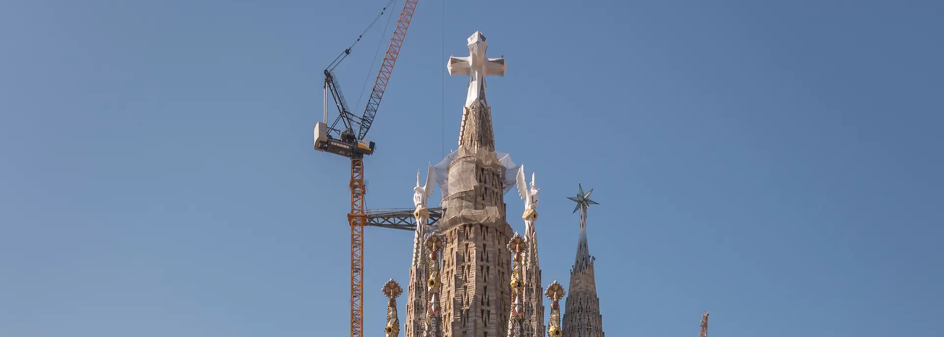 Front view of the Sagrada Familia in Barcelona, with surrounding city buildings in the foreground, construction cranes behind the basilica, and a clear blue sky in the background.