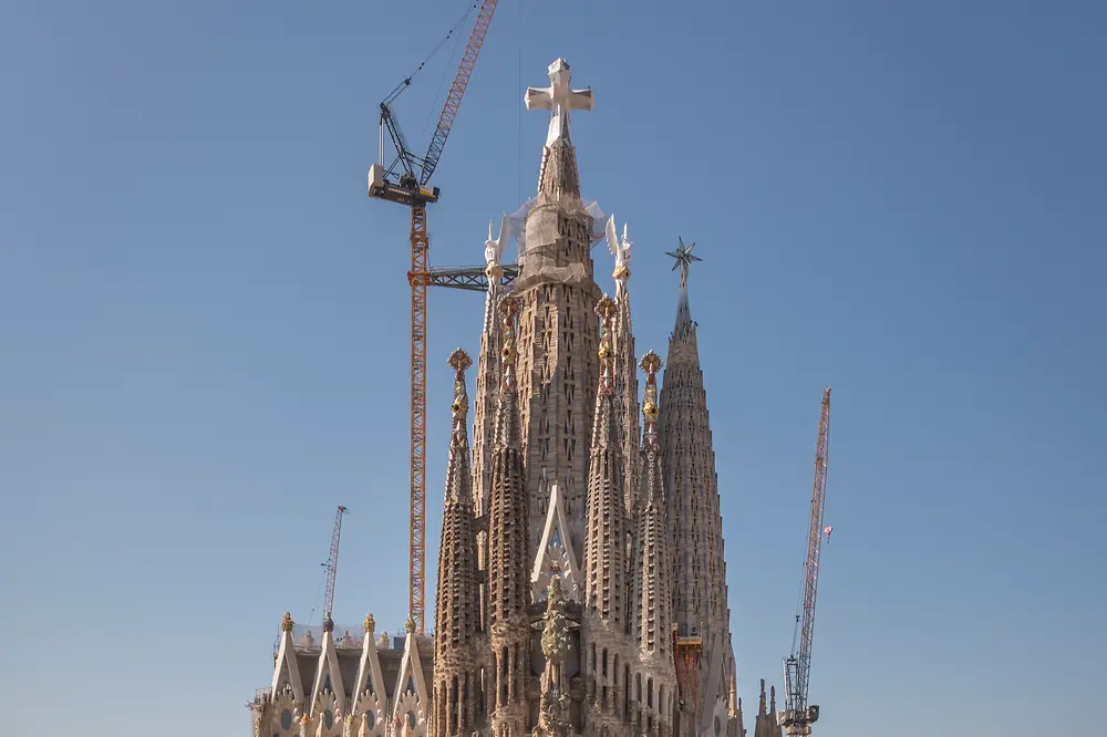 Front view of the Sagrada Familia in Barcelona, with surrounding city buildings in the foreground, construction cranes behind the basilica, and a clear blue sky in the background.