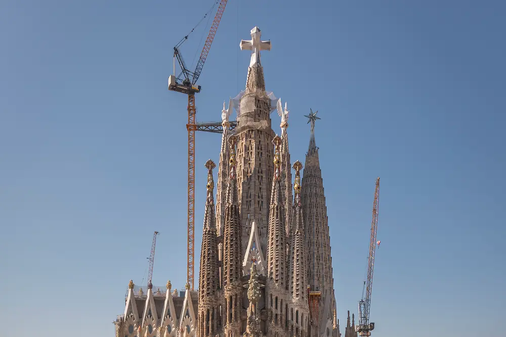 Front view of the Sagrada Familia in Barcelona, with surrounding city buildings in the foreground, construction cranes behind the basilica, and a clear blue sky in the background.